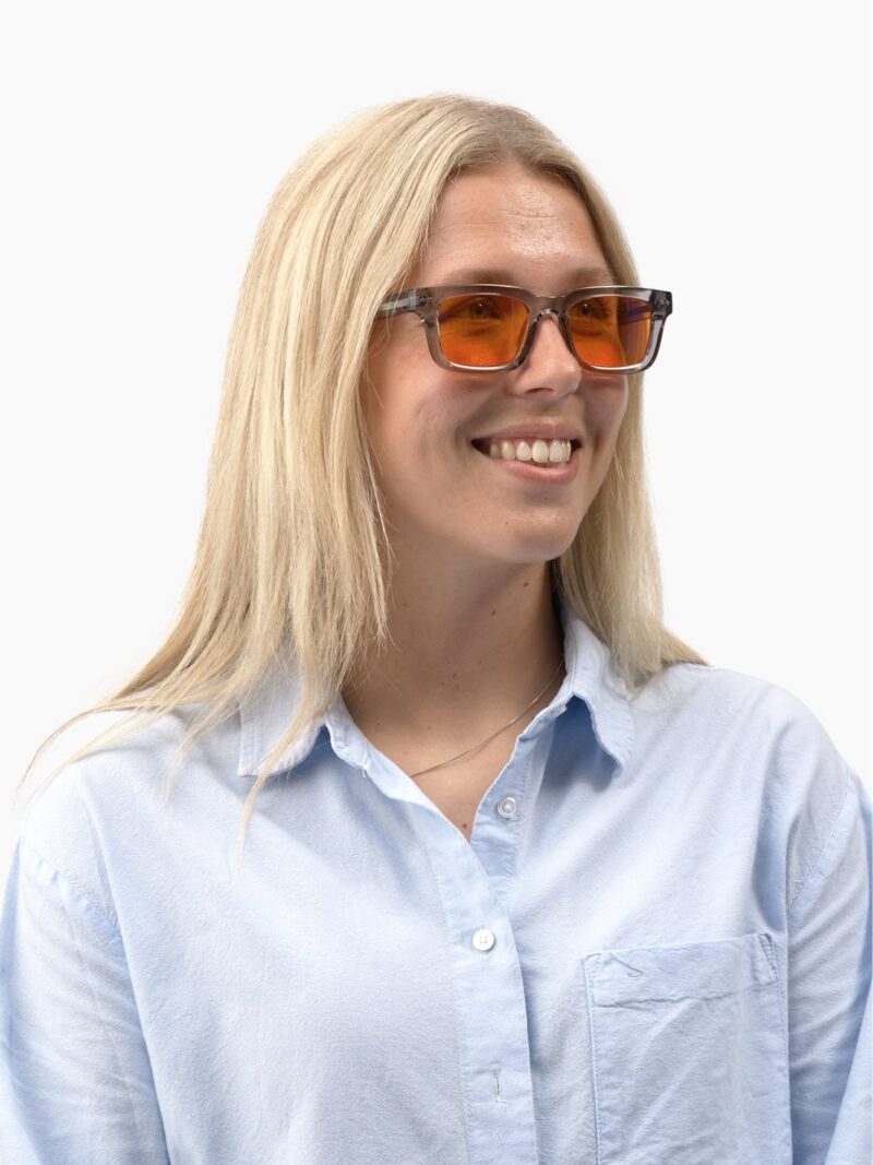 Lächelnde Frau mit langem hellem Haar, der grau-transparenten Eclipse 90 Fassung mit orangefarbenen Gläsern und hellblauem Hemd