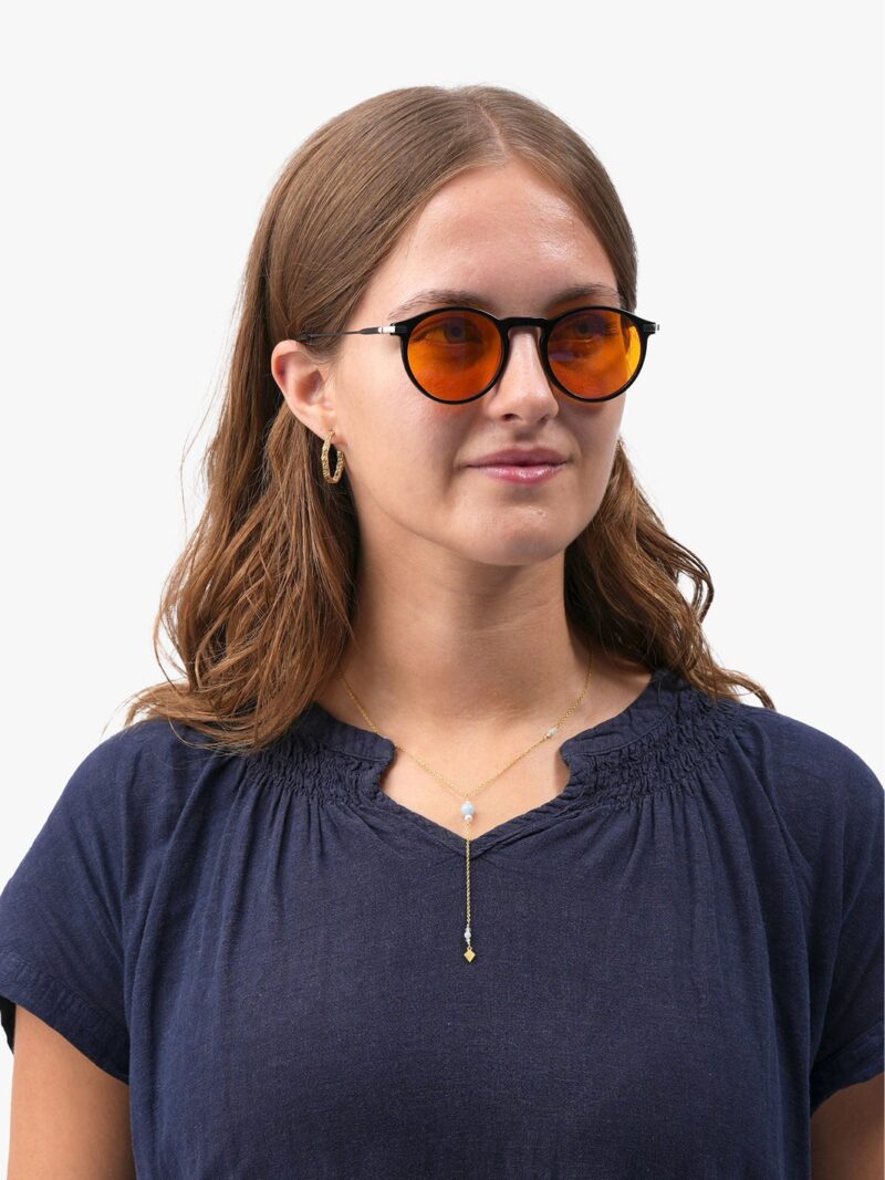 Frau mit runder Brille mit orangefarbenen Gläsern, goldenen Creolen und feiner Lariat-Kette in dunkelblauer Bluse