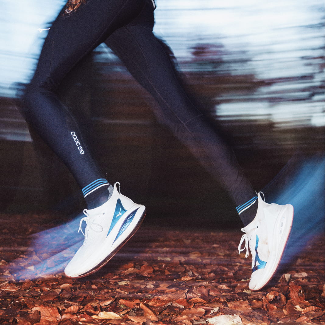 Läufer in Sprungphase, weiße Schuhe mit blauem Akzent, schwarze Leggings, Herbstlaub, Bewegungsunschärfe