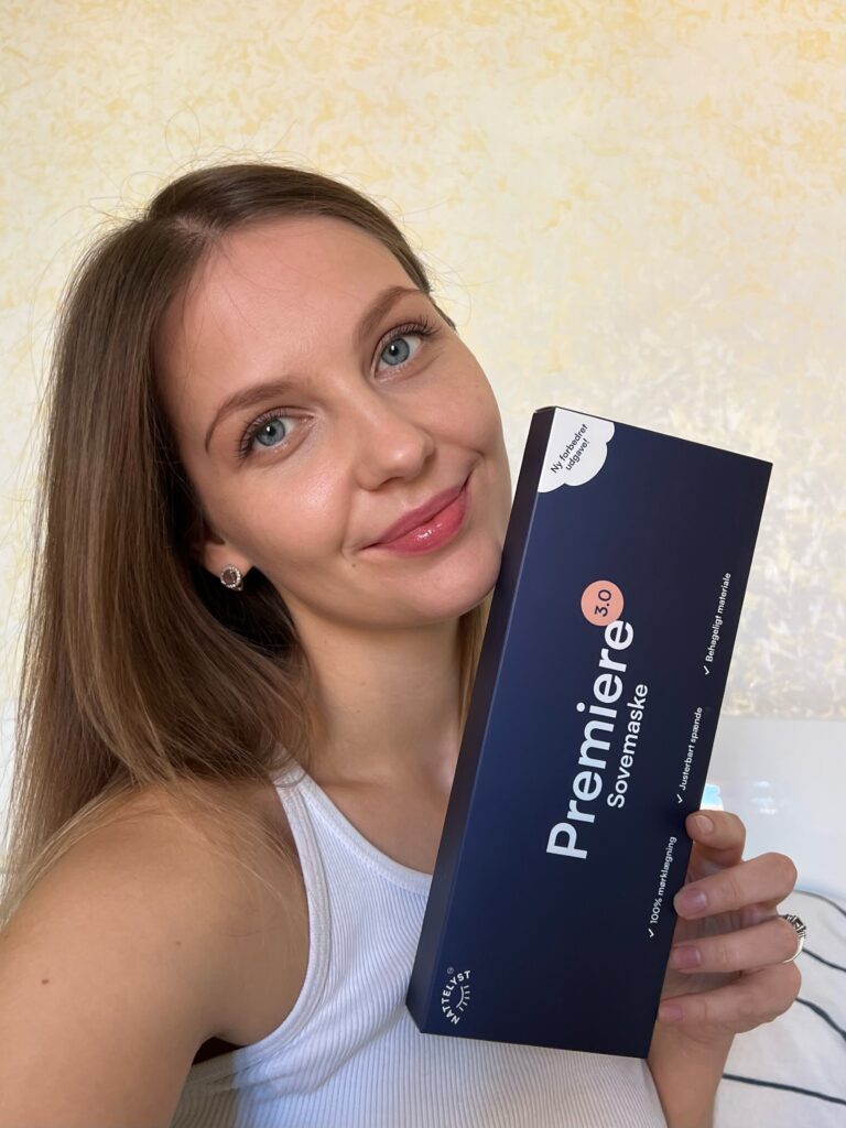 Lächelnde junge Frau mit blauen Augen hält dunkelblaue Verpackung einer Schlafmaske